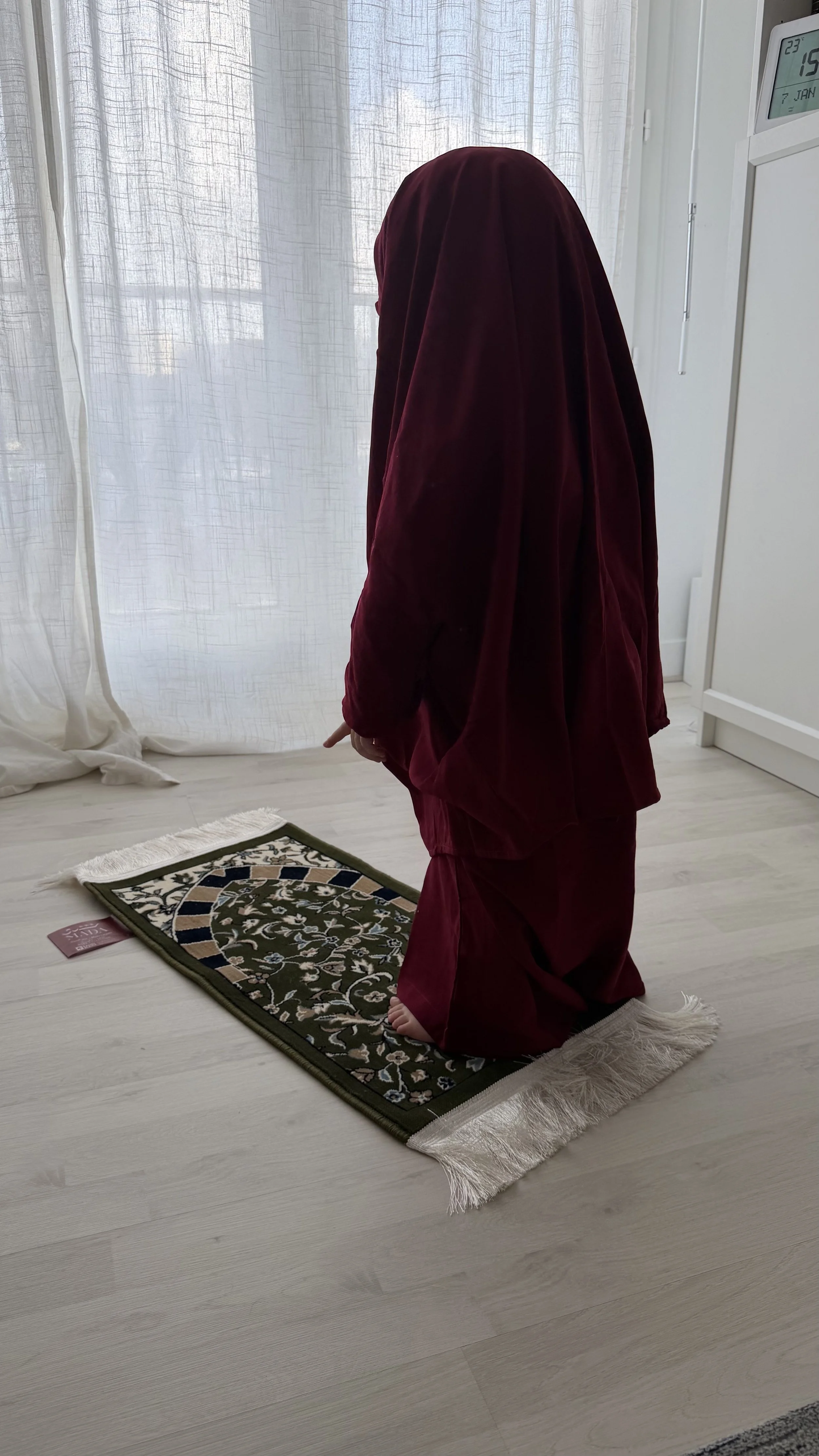 Une personne habillée en vêtements sombres, agenouillée sur un tapis de prière dans une pièce lumineuse avec des rideaux blancs.