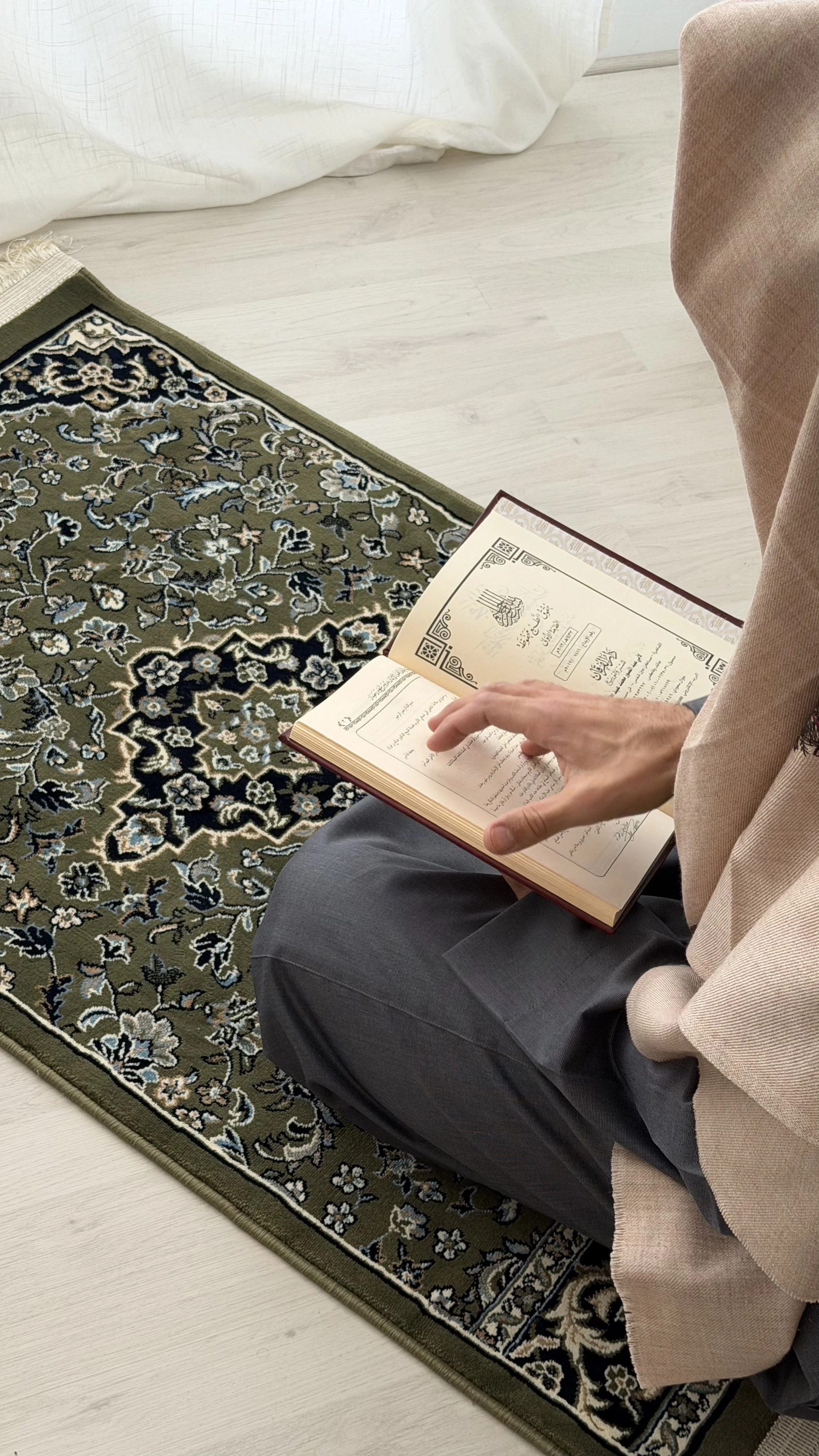 Une personne qui prie ou lit un livre religieux, assise sur un tapis en céramique. La personne porte une veste beige et un pantalon gris. La scène se déroule dans une pièce lumineuse avec un sol en bois clair.
