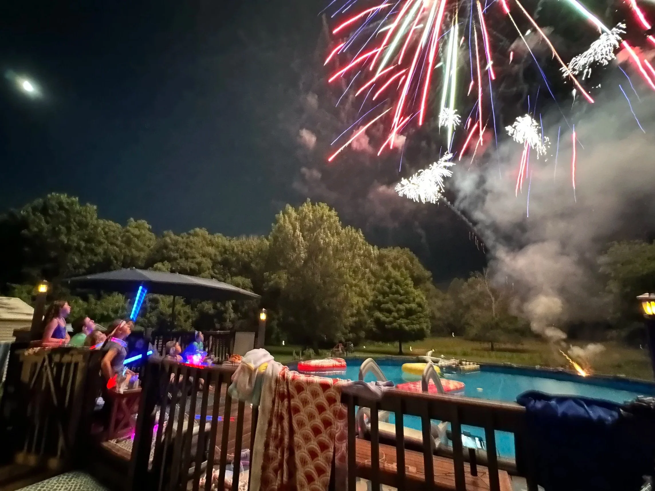 Nighttime scene of a backyard with a pool, children watching fireworks, and trees in the background; fireworks lighting up the sky.