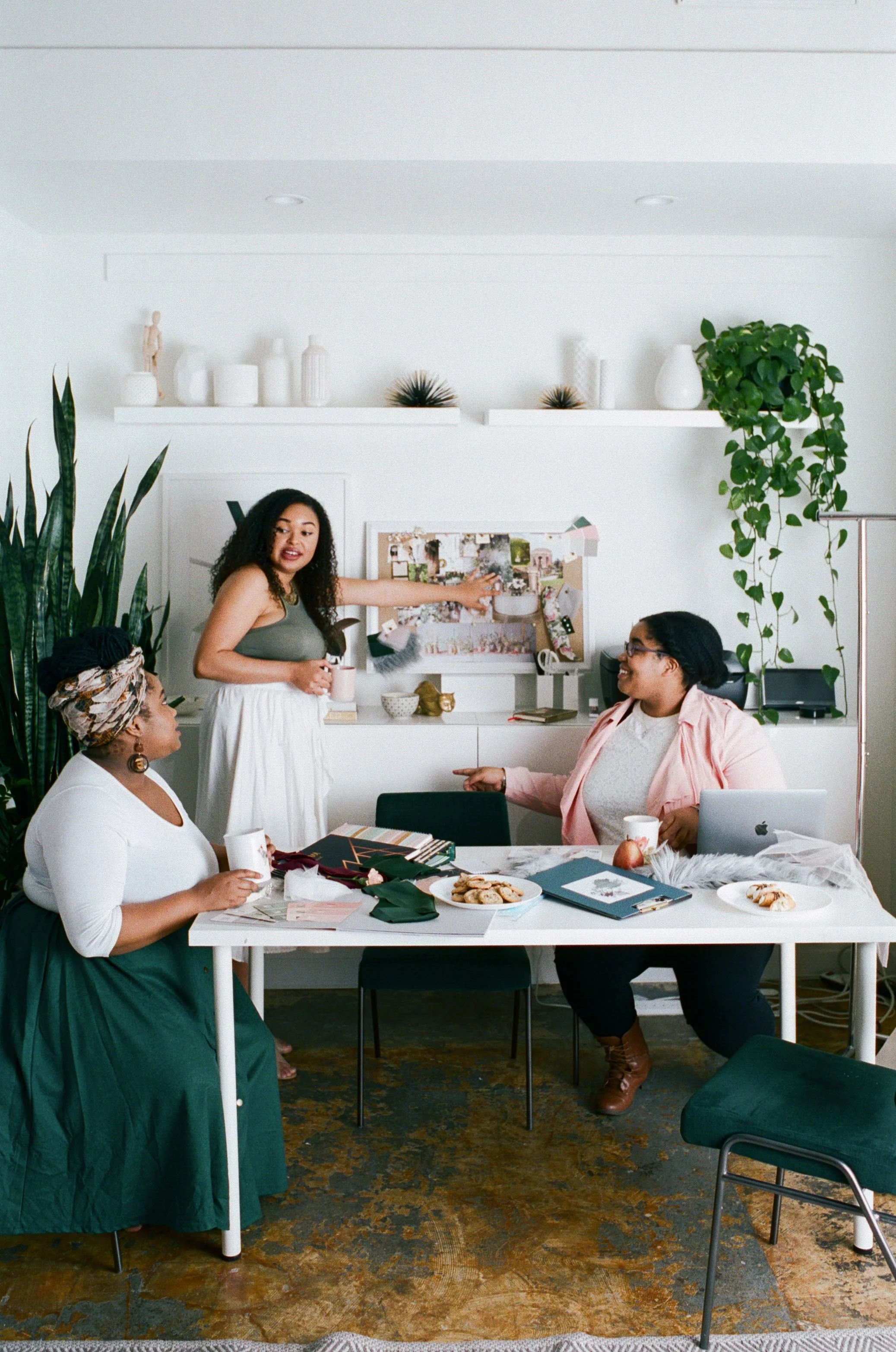 Three women having a discussion in a bright, modern office, with a woman standing and pointing at a bulletin board, while two women sit at a desk with laptops and snacks.