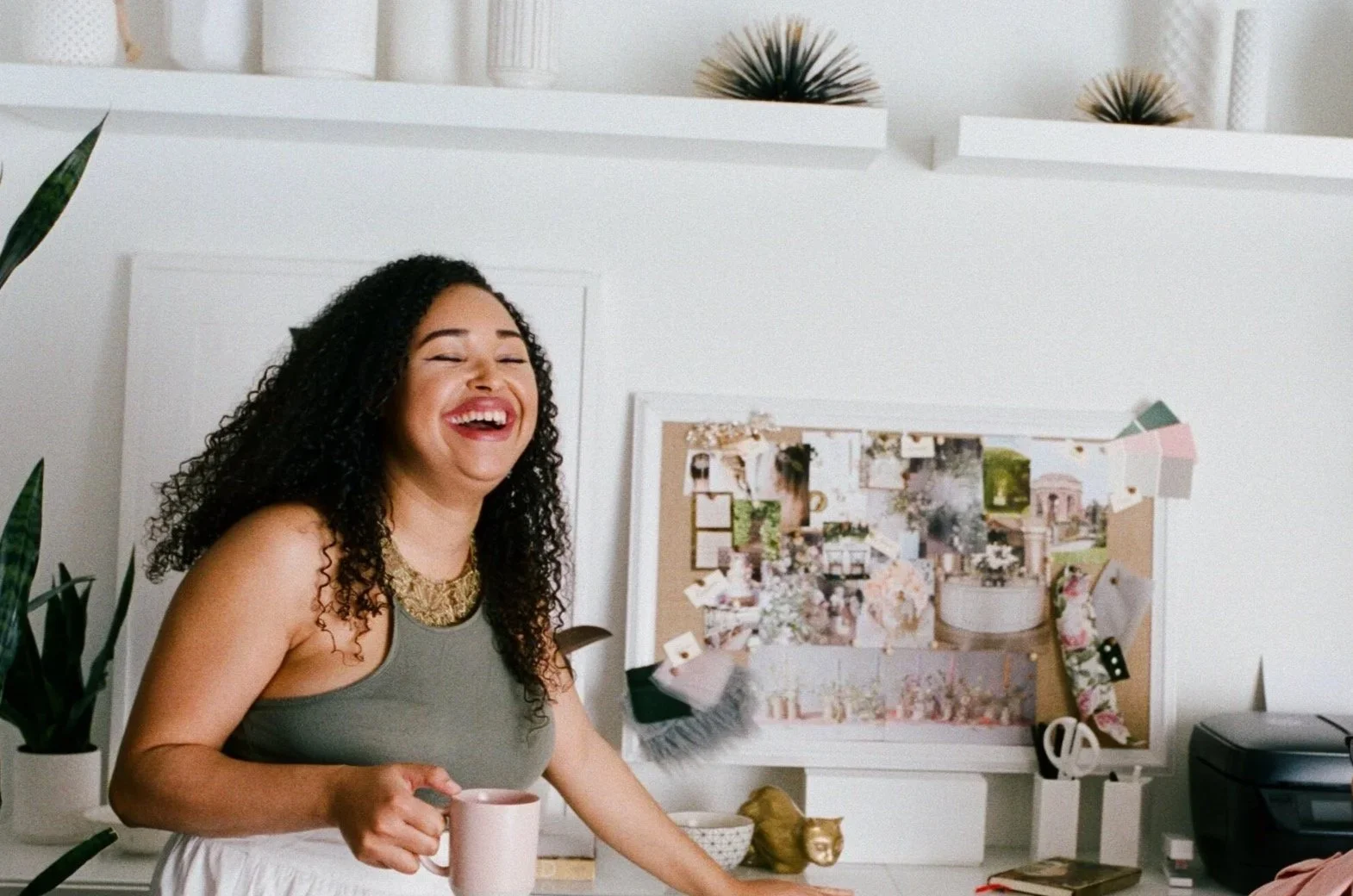 A woman with curly dark hair smiling and laughing, holding a pink mug, standing in a modern, well-decorated room with plants and a bulletin board in the background.