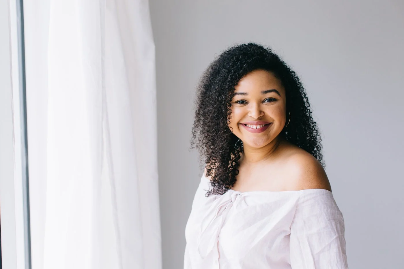 Smiling woman with curly black hair wearing an off-shoulder white top, standing near a window with white curtains.