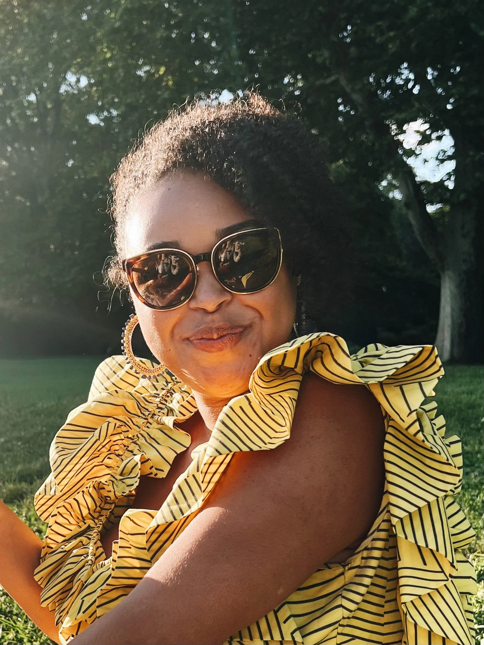 A woman wearing sunglasses and a yellow striped ruffled top sitting outdoors in a grassy area with trees in the background during daylight.