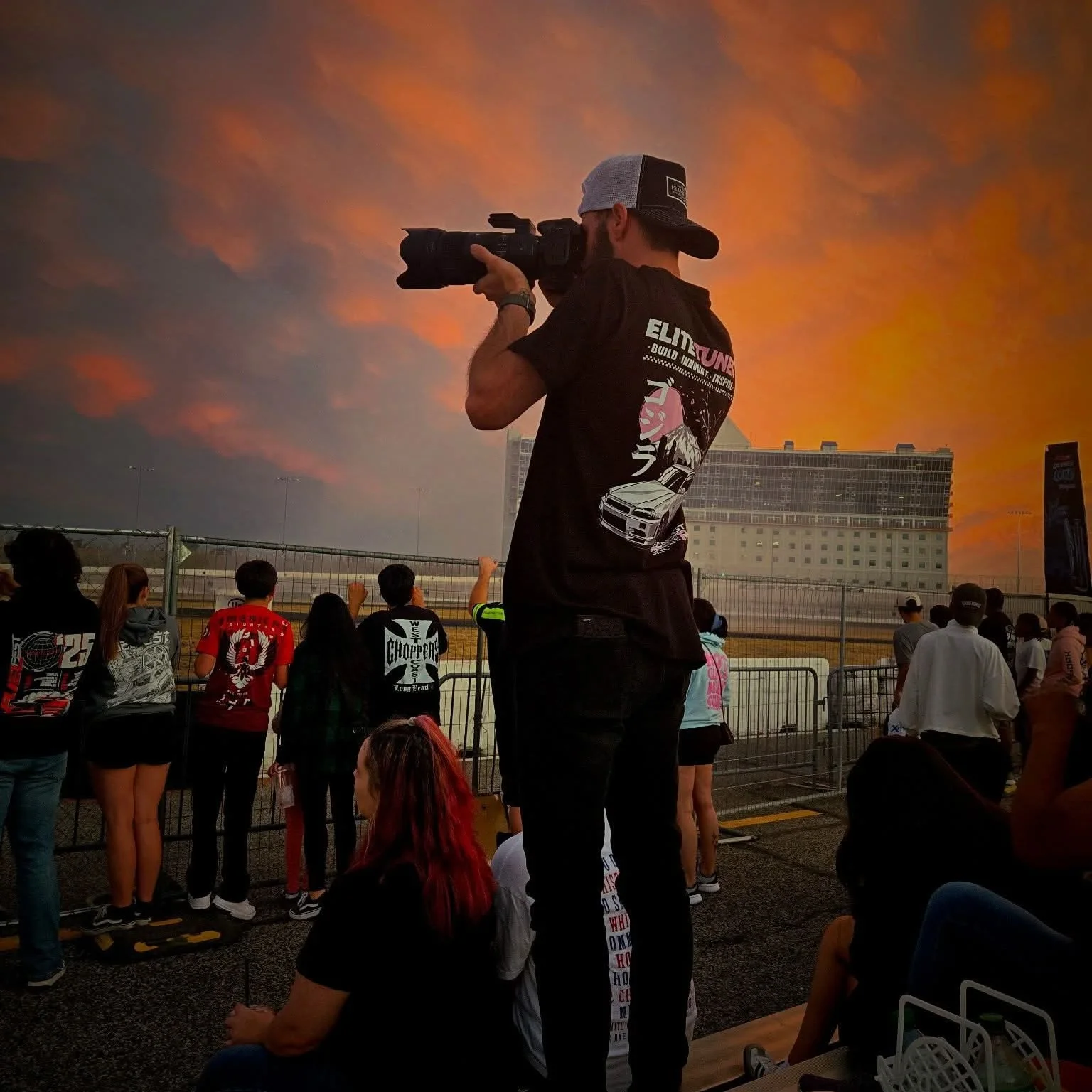 A photographer with a camera on his shoulder capturing a race track event with a sunset sky in the background, spectators watching behind a fence.