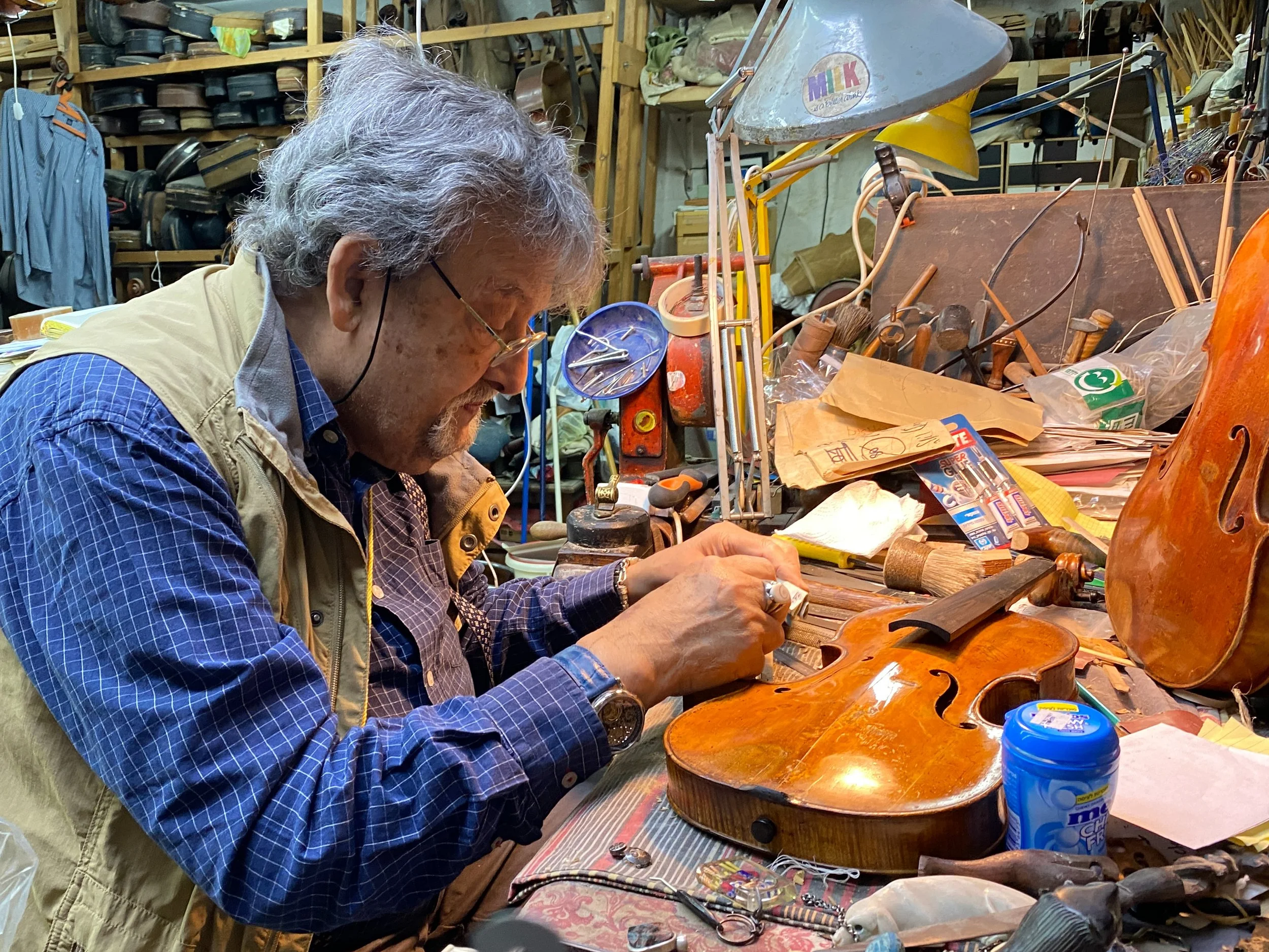 An older man working on a violin at a cluttered woodworking bench in a workshop, surrounded by tools and wood pieces.