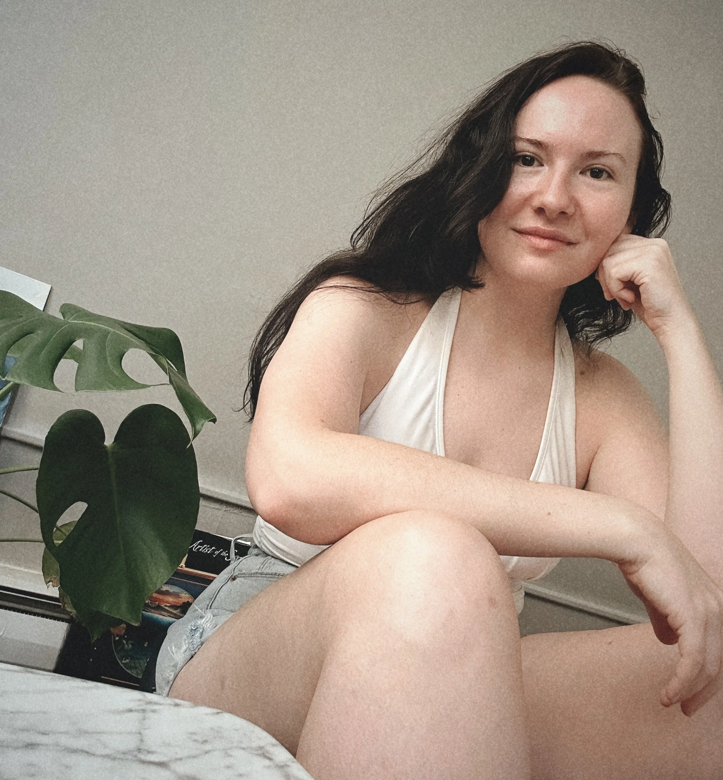 Rachel Leedom, a young woman with black hair and fair skin sitting indoors, wearing a white top and distressed denim shorts, with a large green plant and a bookshelf in the background.