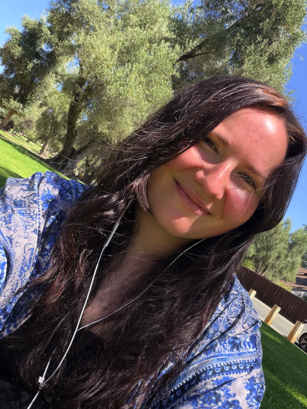 A young woman with long, dark, wavy hair taking a selfie outdoors on a sunny day, smiling softly. She is wearing a blue patterned shirt and has white earphones. The background features green trees, grass, and a clear blue sky.