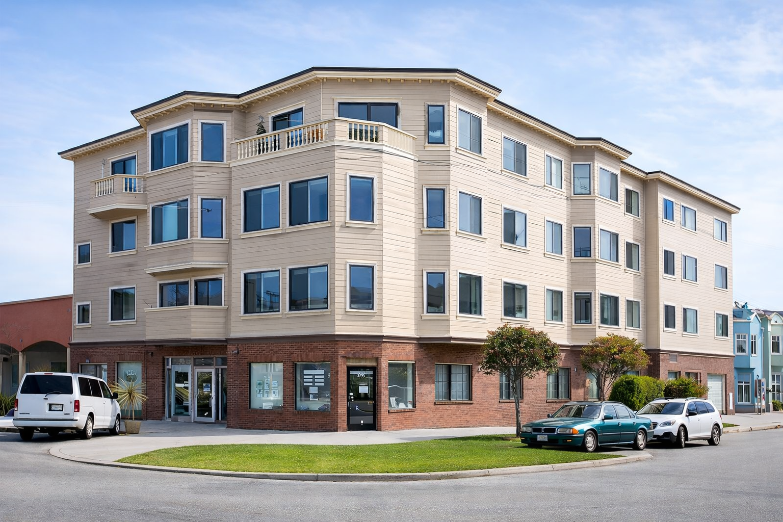 A multi-story residential building with beige siding and a red brick base, featuring many windows, with cars parked outside and small trees along the sidewalk.