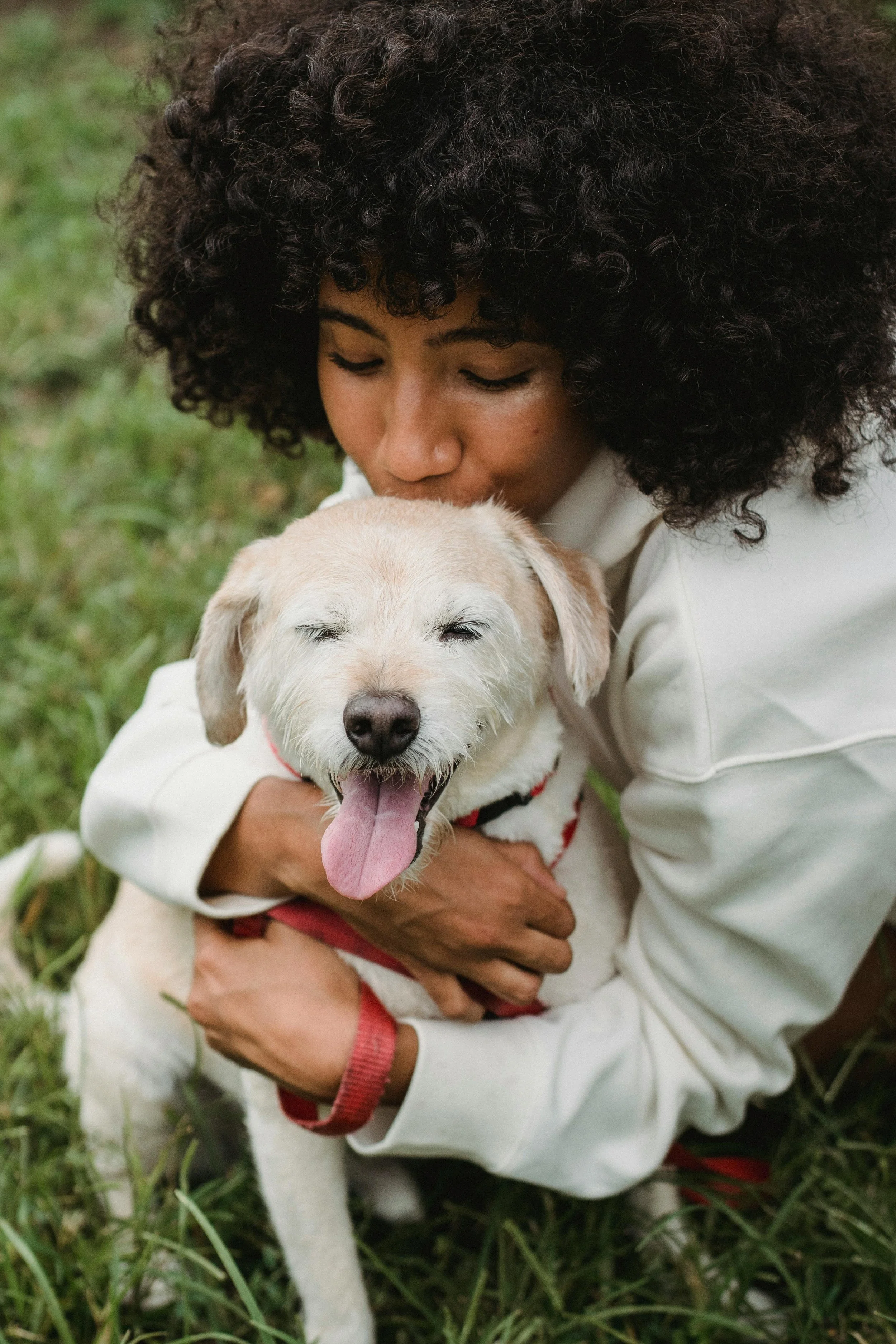 A woman with curly hair hugging a smiling dog with closed eyes outdoors on grass.