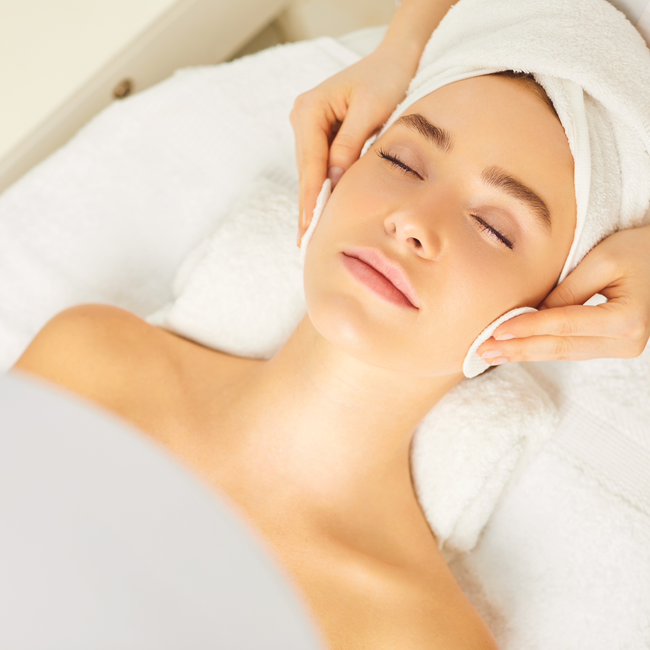 Woman receiving a facial massage with a towel wrapped around her head, lying peacefully with eyes closed.