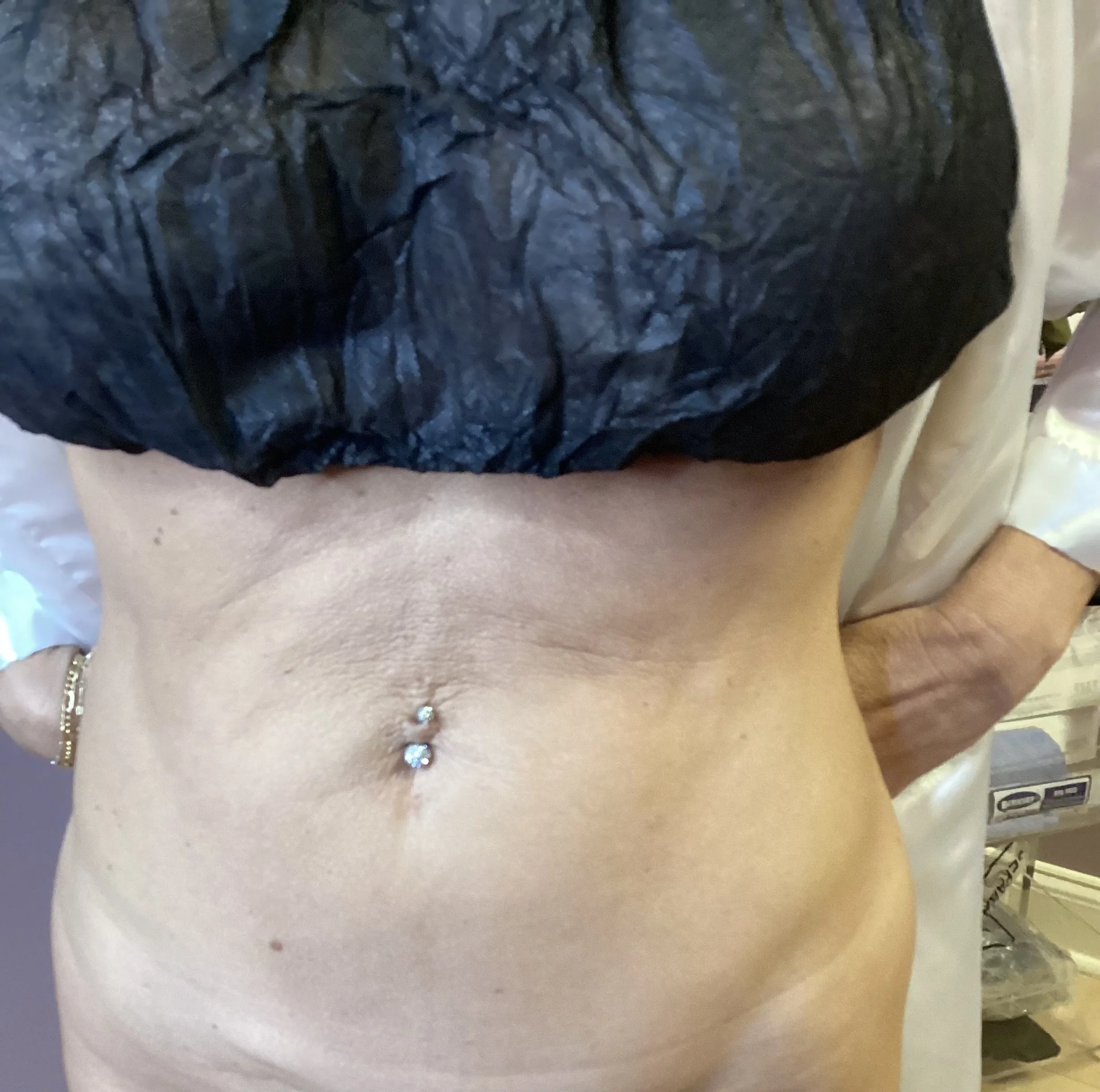 Close-up of a woman's midsection, showing her belly button with a diamond stud piercing, wearing a black upper garment and gold earrings.