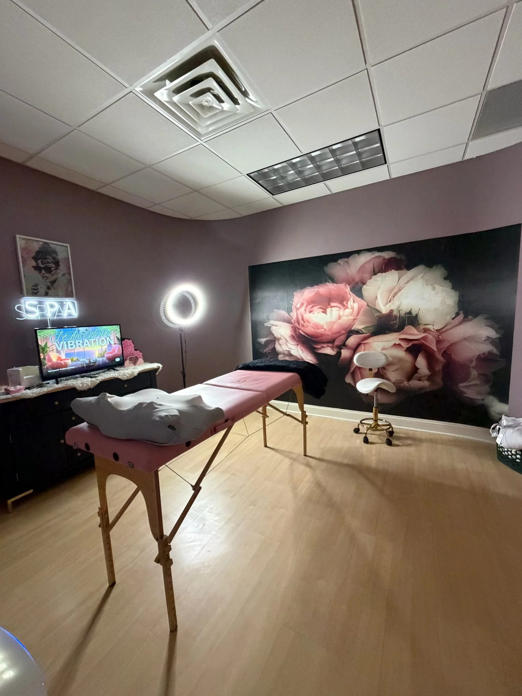 An interior room with a massage table, a large floral wall mural, a neon sign, a television, and decorative lighting.