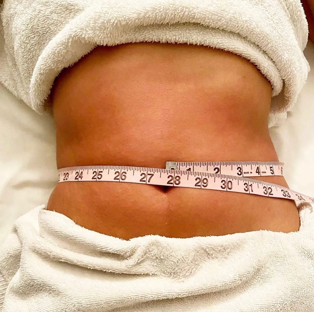 Close-up of a person's midsection with a measuring tape around their waist, showing a measurement of approximately 28 inches. The person is wearing a white towel or robe.