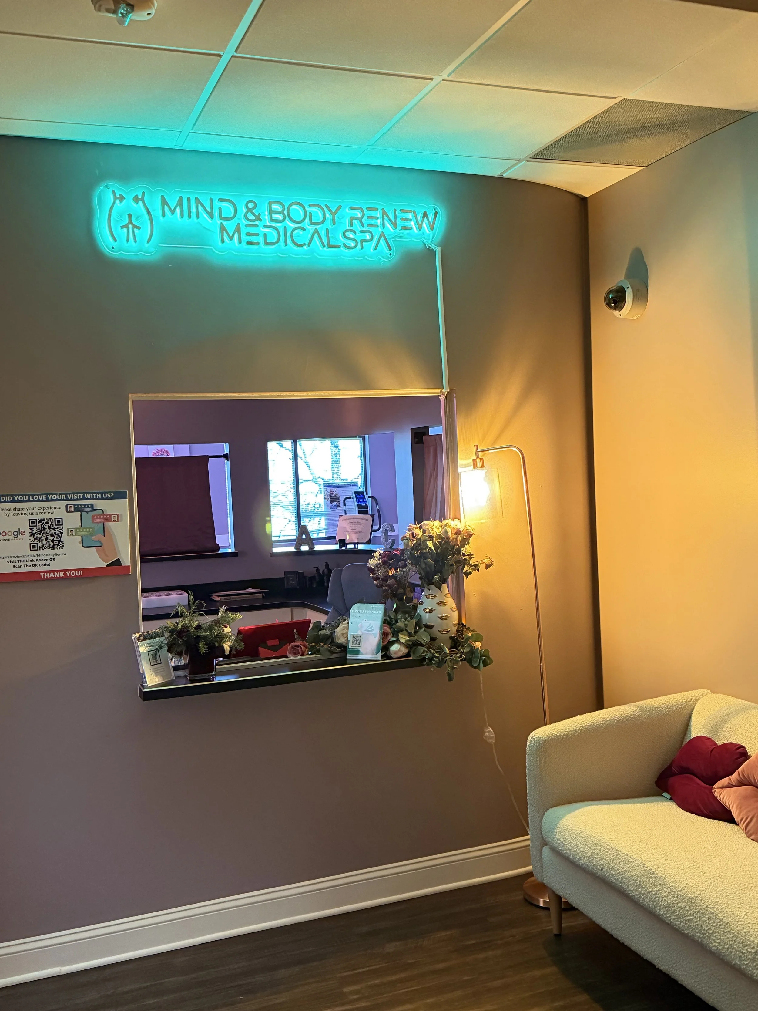 Interior of a medical spa with a neon sign that reads 'Mind & Body Renew Medical Spa', a mirror above a small window, a couch with pillows, a floor lamp, and a table with flowers.