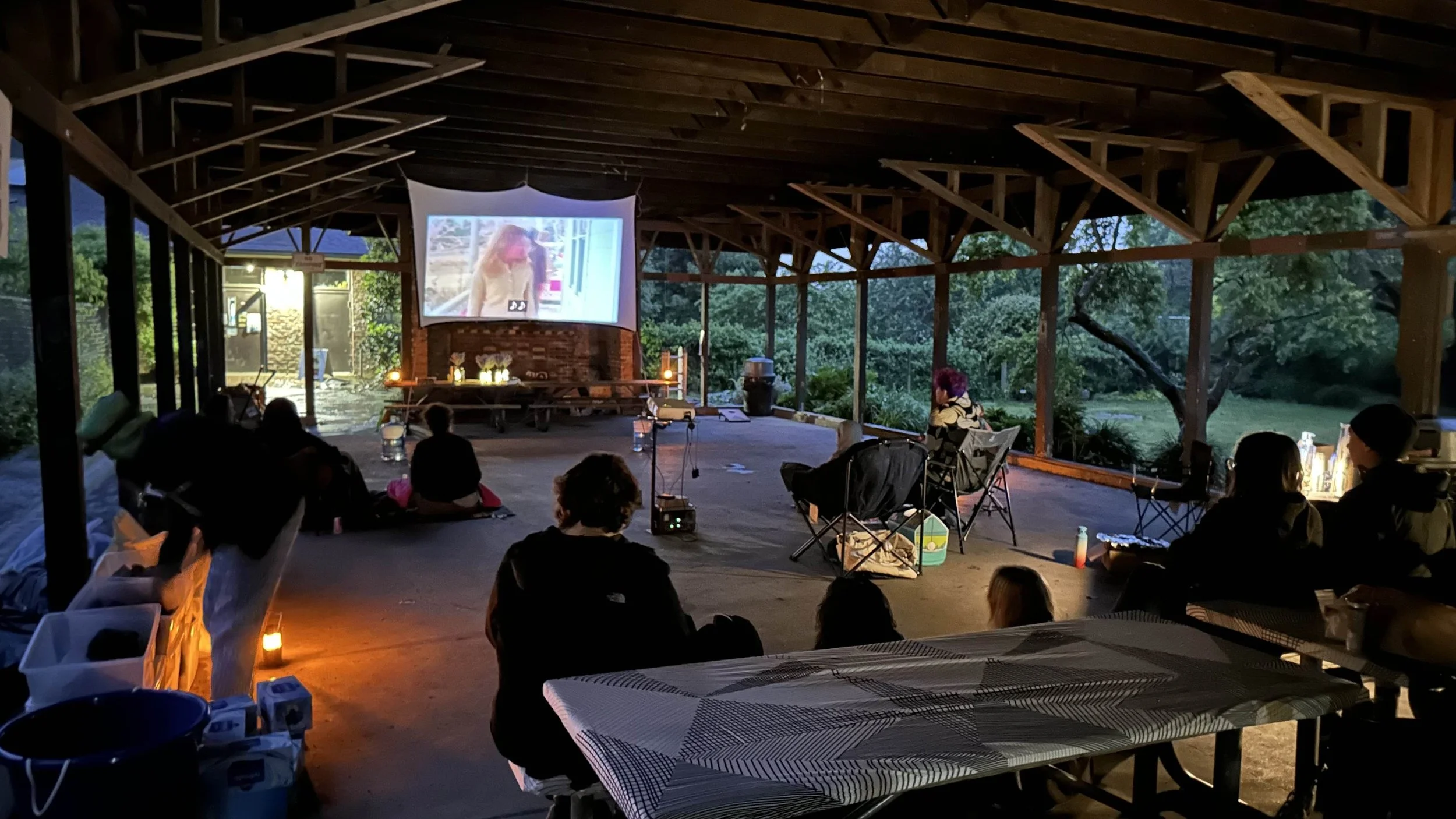 People gathered inside a large open-air pavilion watching a movie projected on a screen, with trees visible outside.