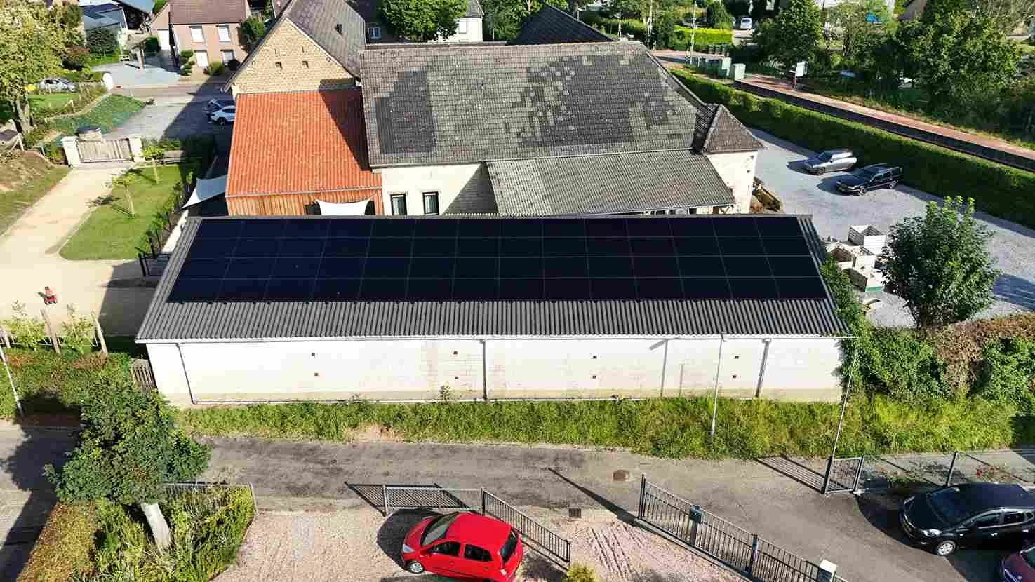 Luchtfoto van een gebouw met zonnepanelen op het dak, omringd door huizen, een rode auto en groen.