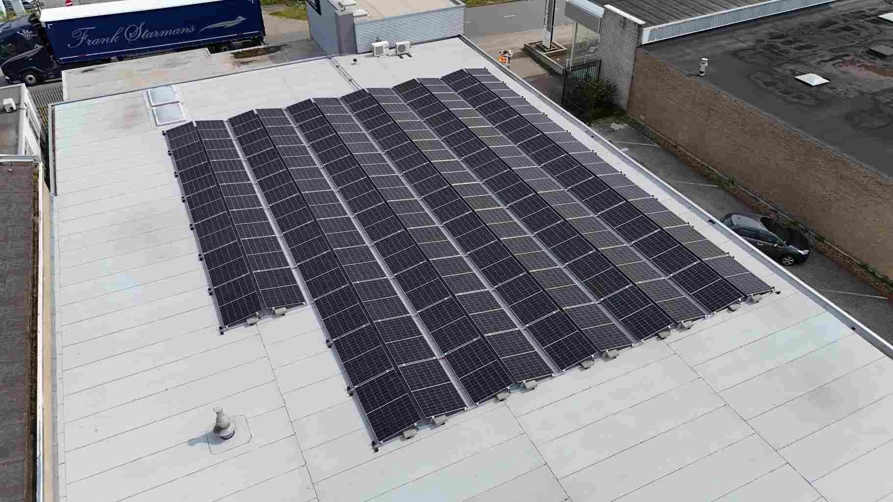 Zonnepanelen op een plat dak van een gebouw, met een vrachtwagen en auto op de achtergrond.