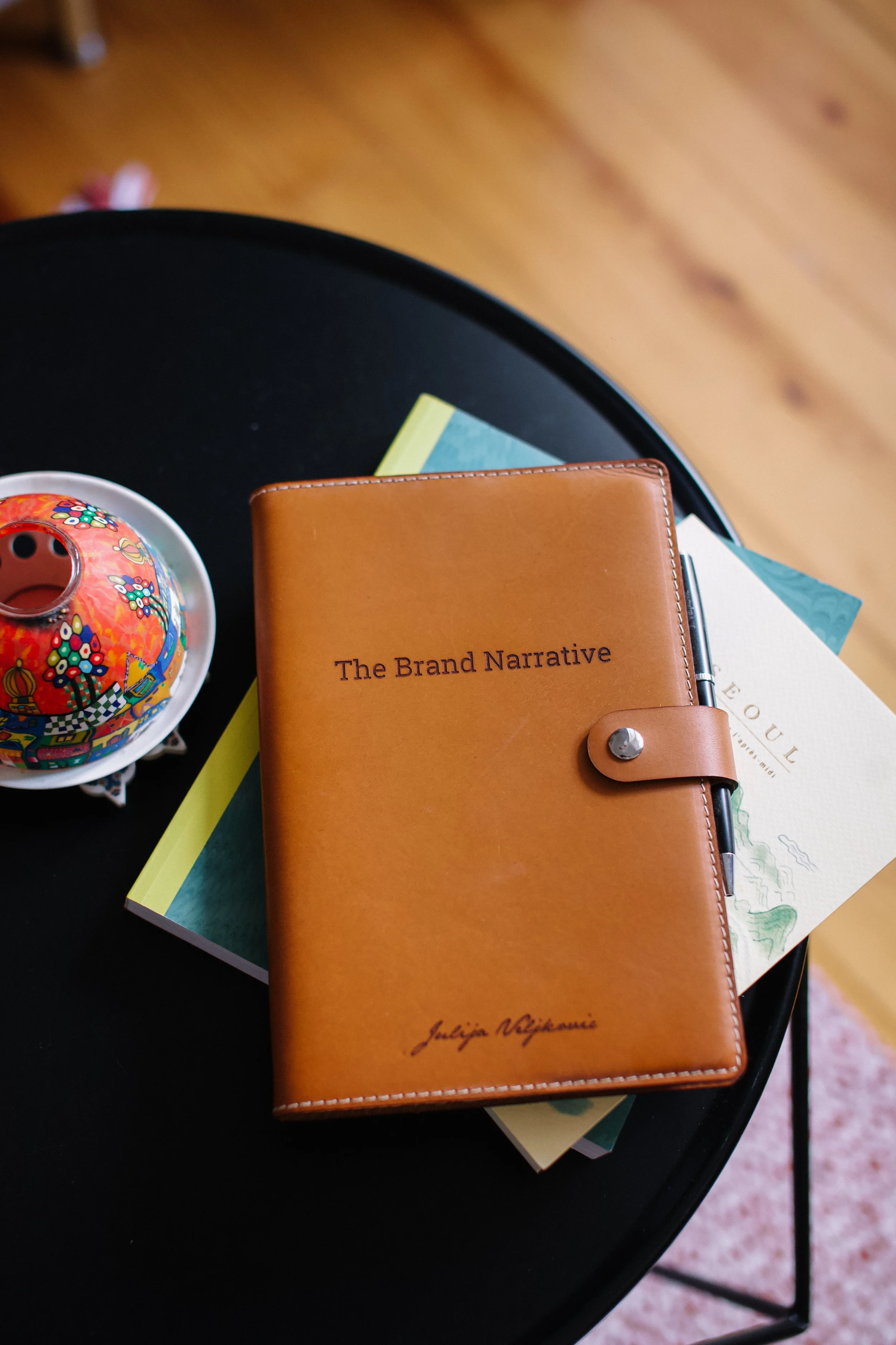 A calendar, a leather-bound notebook titled 'The Brand Narrative', and a colorful decorative item on a black round table.
