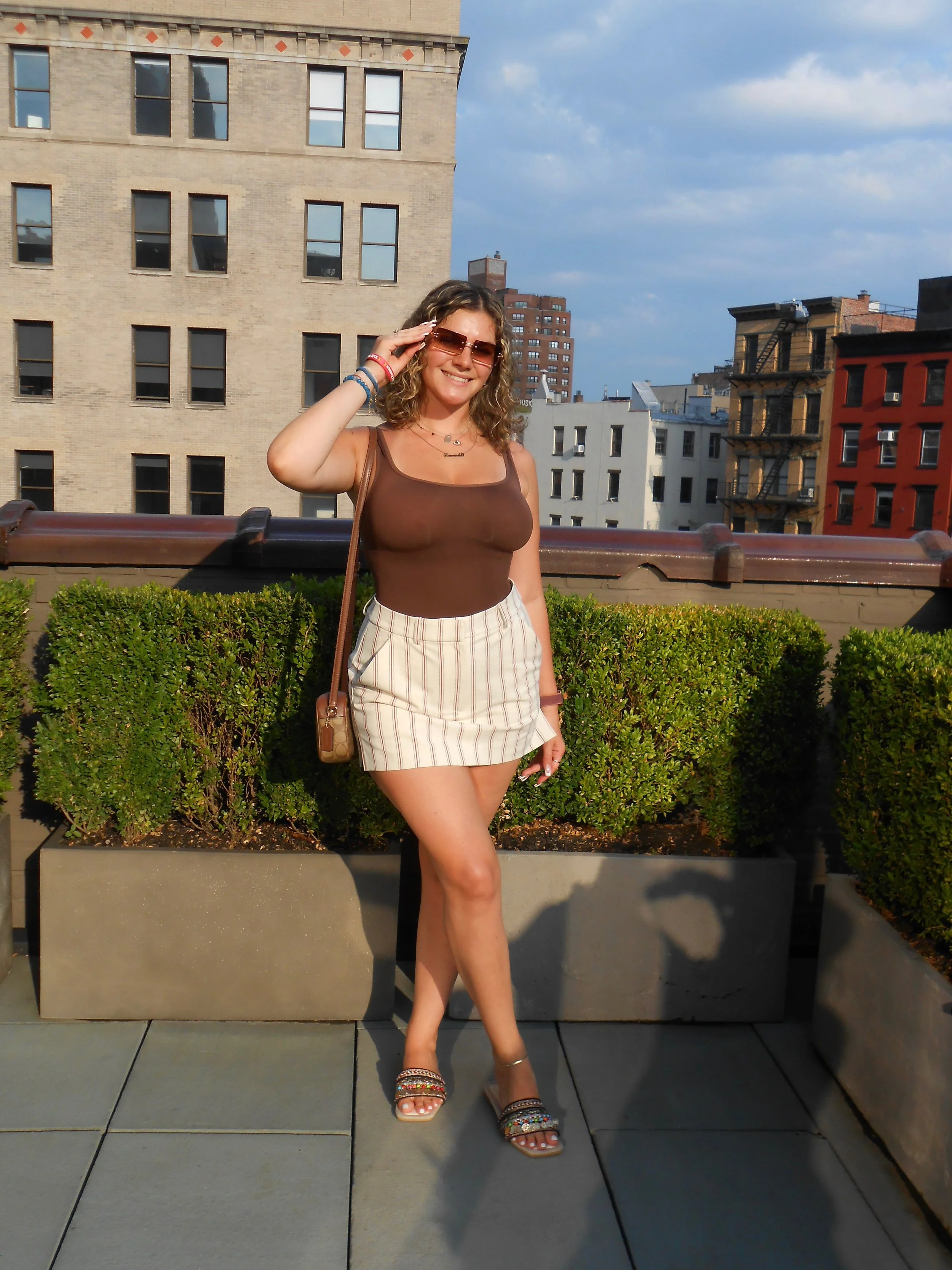 A woman with curly hair wearing sunglasses, a sleeveless brown top, a striped beige skirt, and open-toed sandals, standing on a rooftop with city buildings in the background, smiling and adjusting her sunglasses.