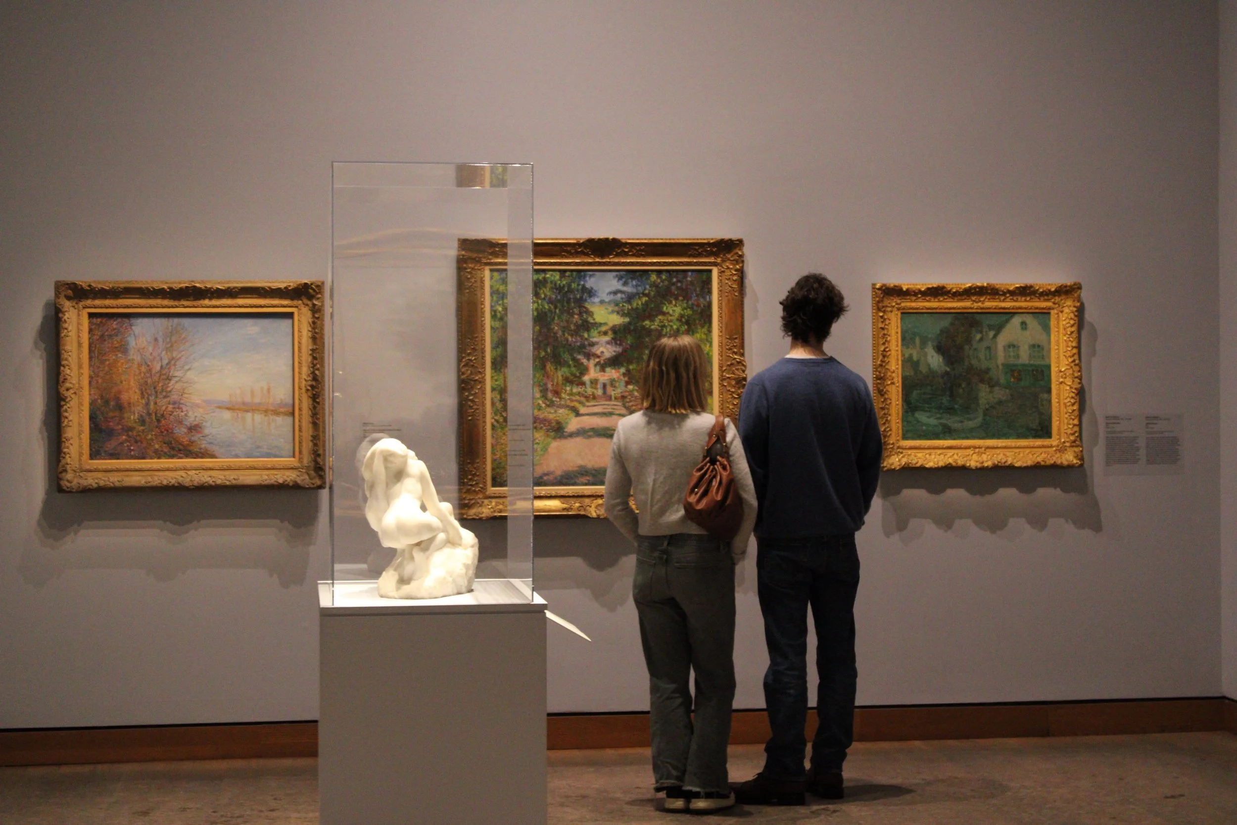 Two people, a woman and a man, stand in front of framed landscape paintings in an art museum, facing the wall, looking at the artwork.