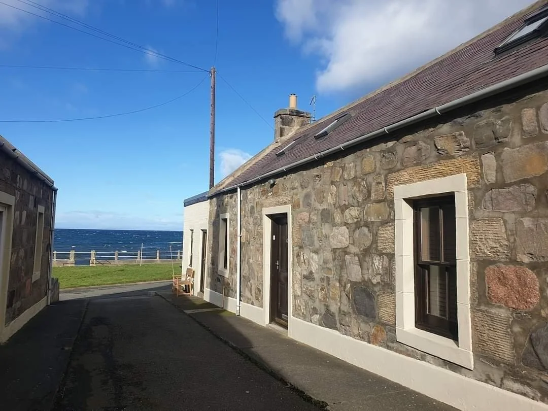 A narrow alley between stone houses with a view of the ocean in the distance, under a partly cloudy blue sky.