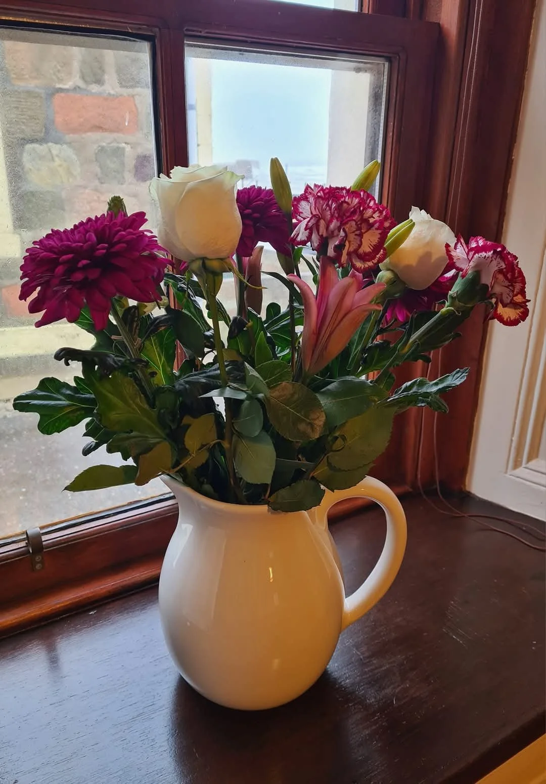A white ceramic pitcher filled with a bouquet of various flowers, including white roses, pink lilies, and purple flowers, placed on a wooden surface by a window.
