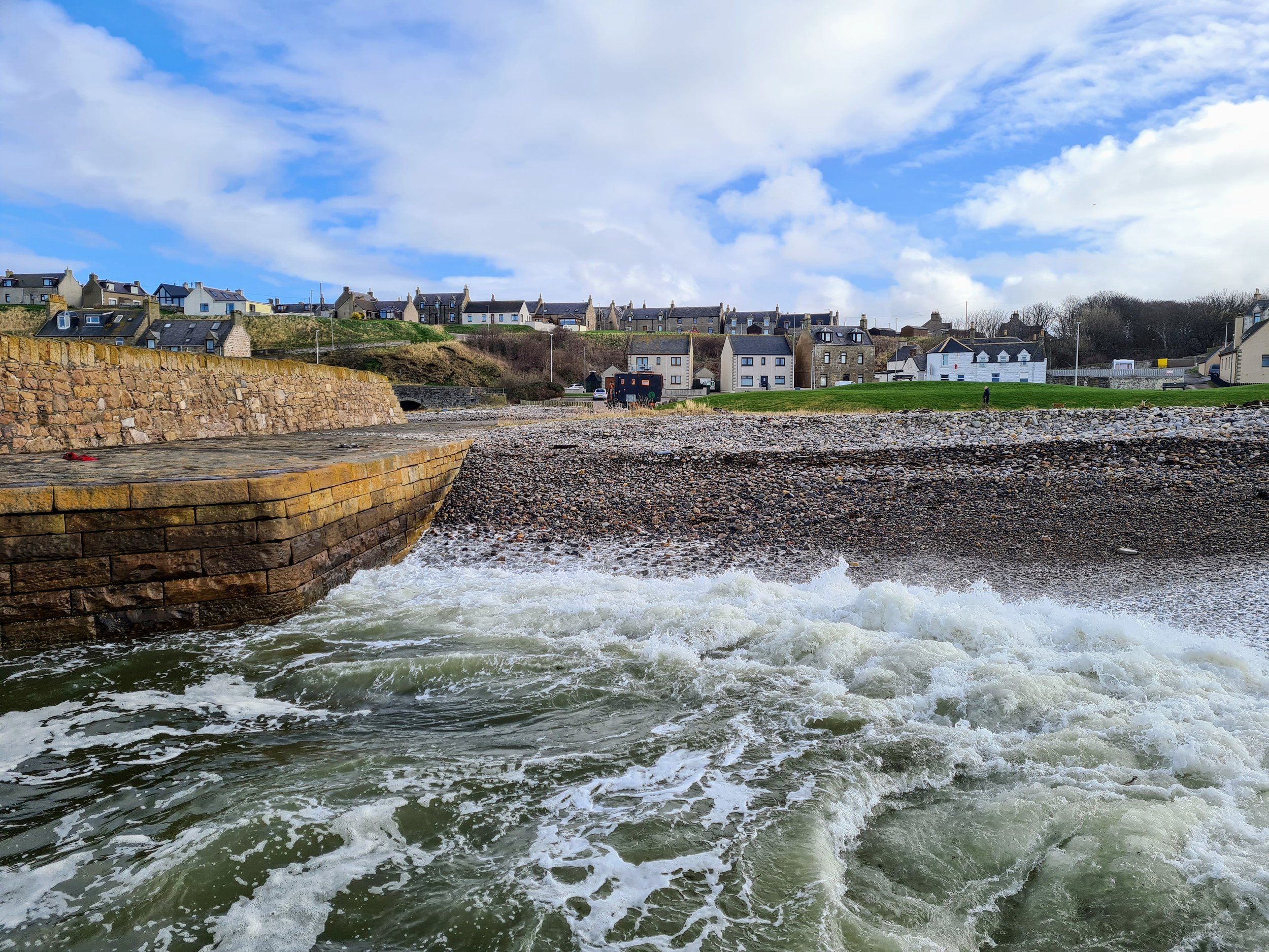 View of water flowing over a small dam with houses and buildings on a hill in the background under a partly cloudy sky.