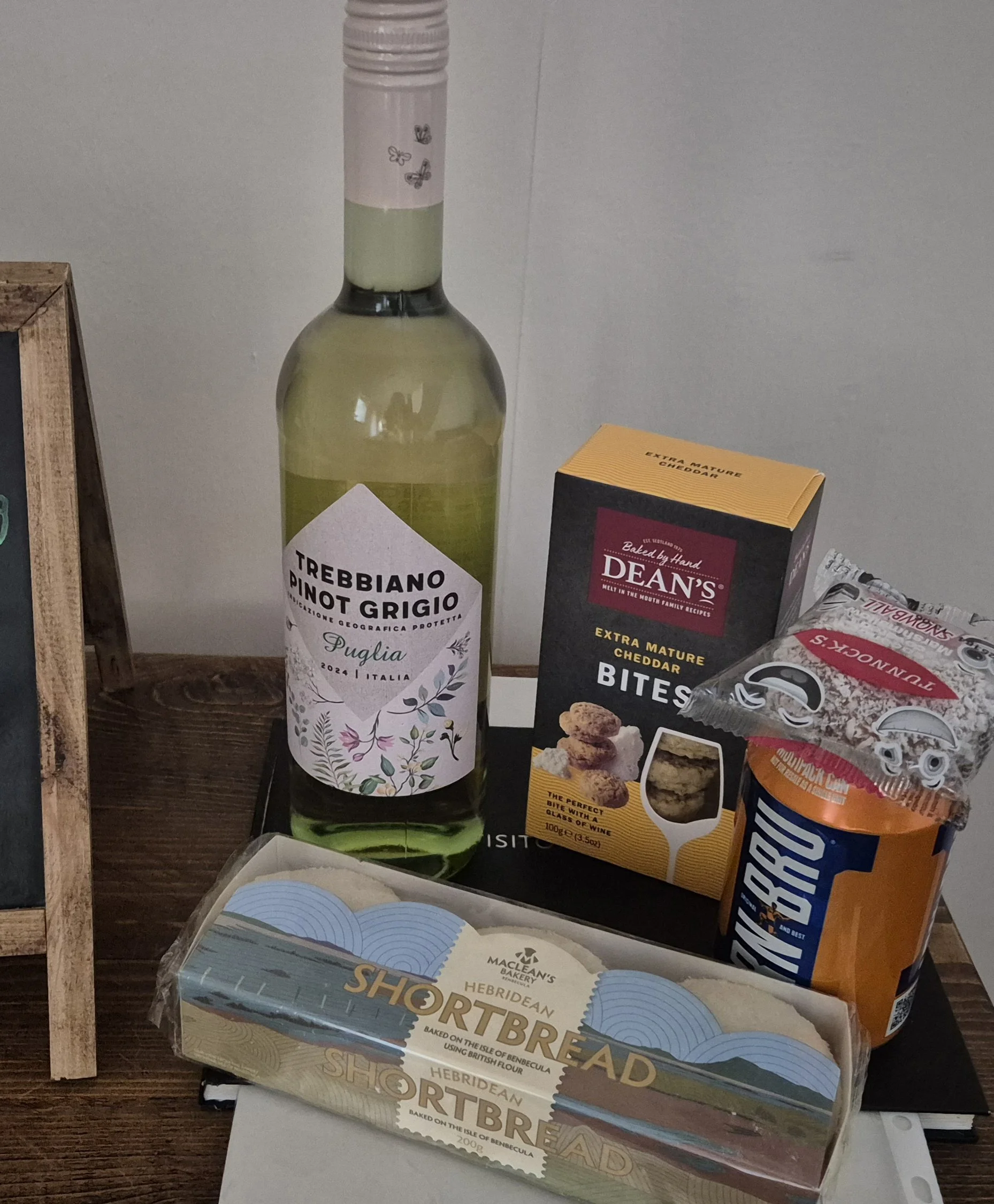 A bottle of Trebbiano Pinot Grigio wine, a box of Dean's extra mature cheddar bites, a packet of Snickers Almond chocolate bar, and a package of Maclean's Shetland Shortbread baked on the Isle of Bencuela.
