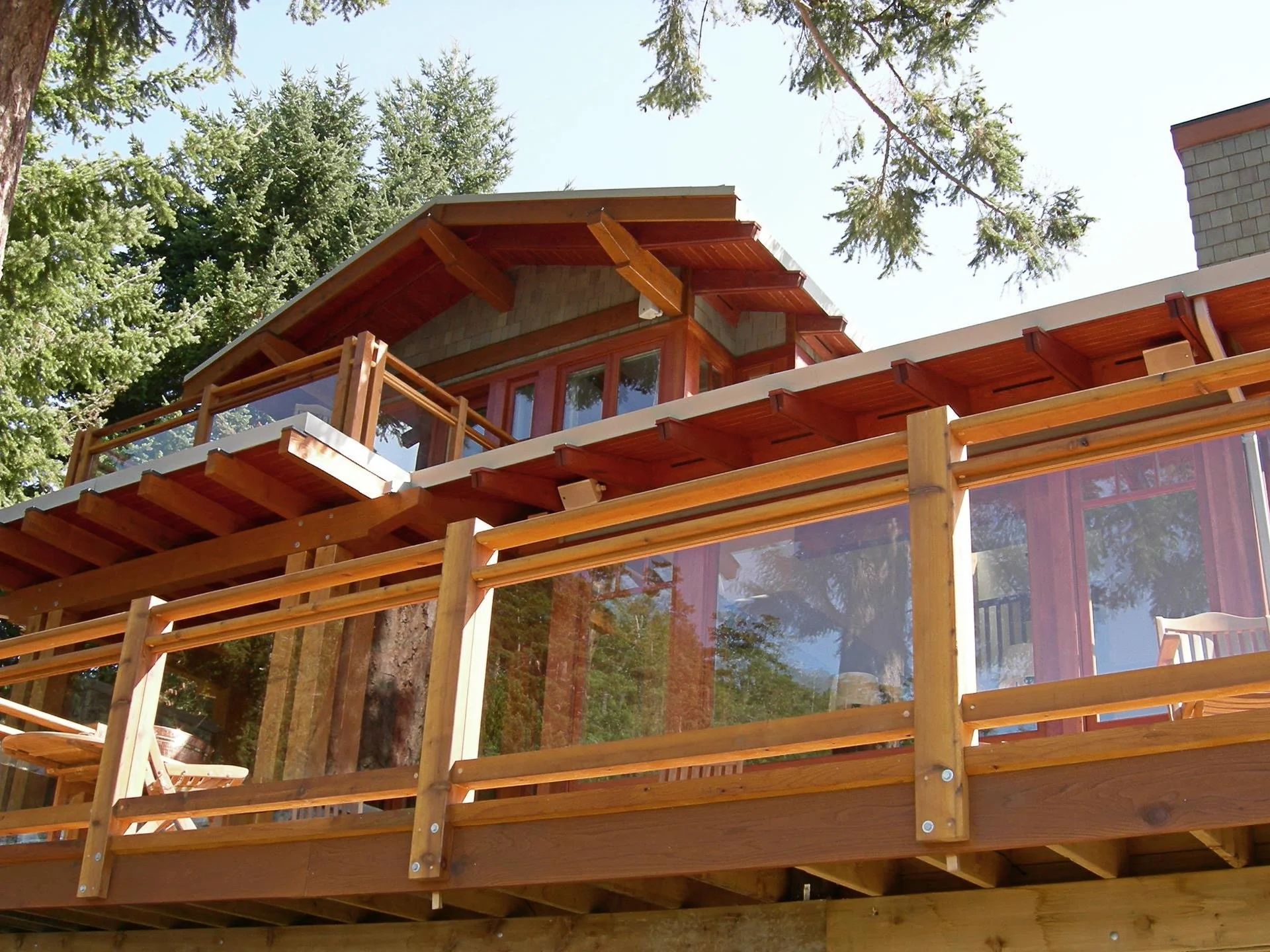 A multi-level wooden house with large glass windows, surrounded by pine trees, with a prominent wooden deck and balcony built with wooden railings.