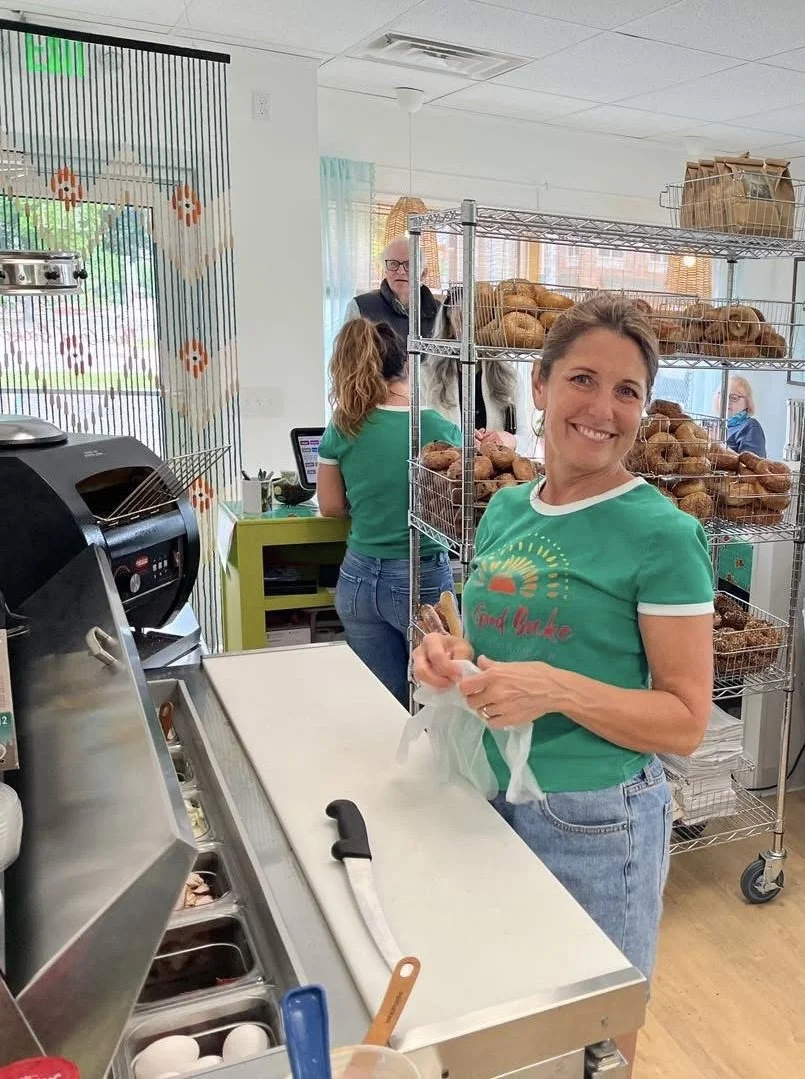 A woman smiling and holding a plastic bag inside a bakery or cafe, with a metal rack of assorted baked goods behind her. Other people are in the background, with a man at the counter and a woman working on a laptop.