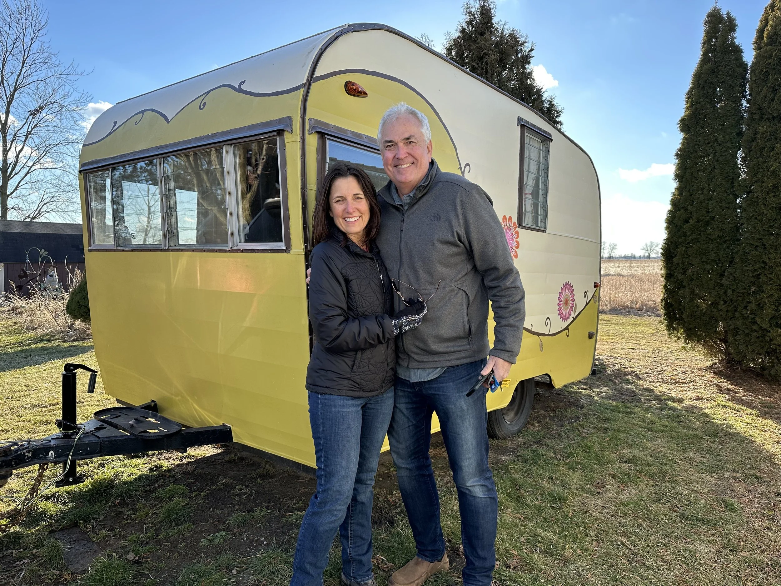 A man and woman smiling, standing outside near a painted yellow vintage trailer with colorful flower designs, on a sunny day with a clear sky and trees in the background.