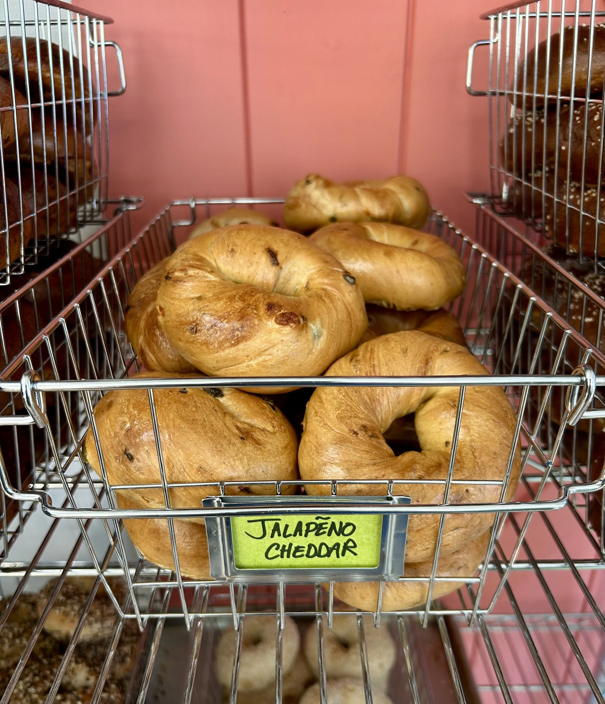 Bakery basket filled with 'Jalapeño Cheddar' bagels against a pink background.