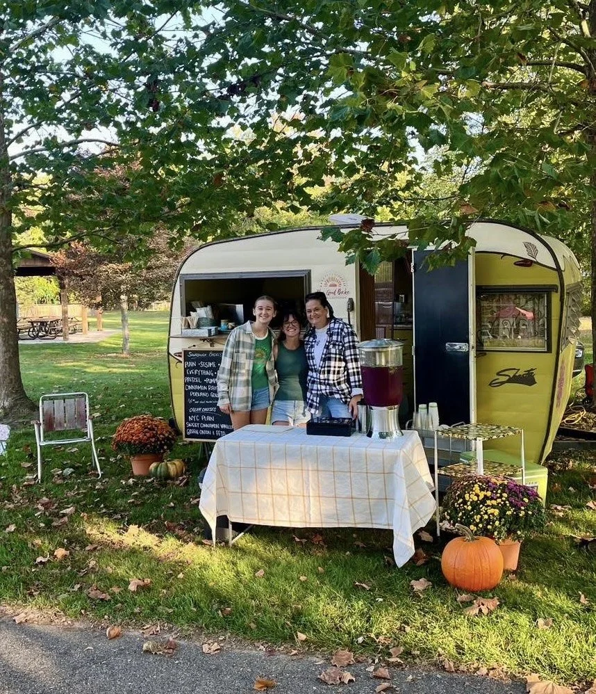 Three women standing in front of a vintage trailer used as a small food or beverage stand, with a table covered by a checkered cloth and decorated with pumpkins and flowers. The setting is outdoors in a park surrounded by trees with autumn leaves.