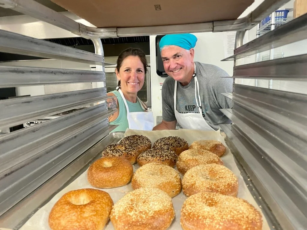 Two people, a woman and a man wearing a headscarf, pose behind a tray of assorted bagels on a baking sheet inside an oven.
