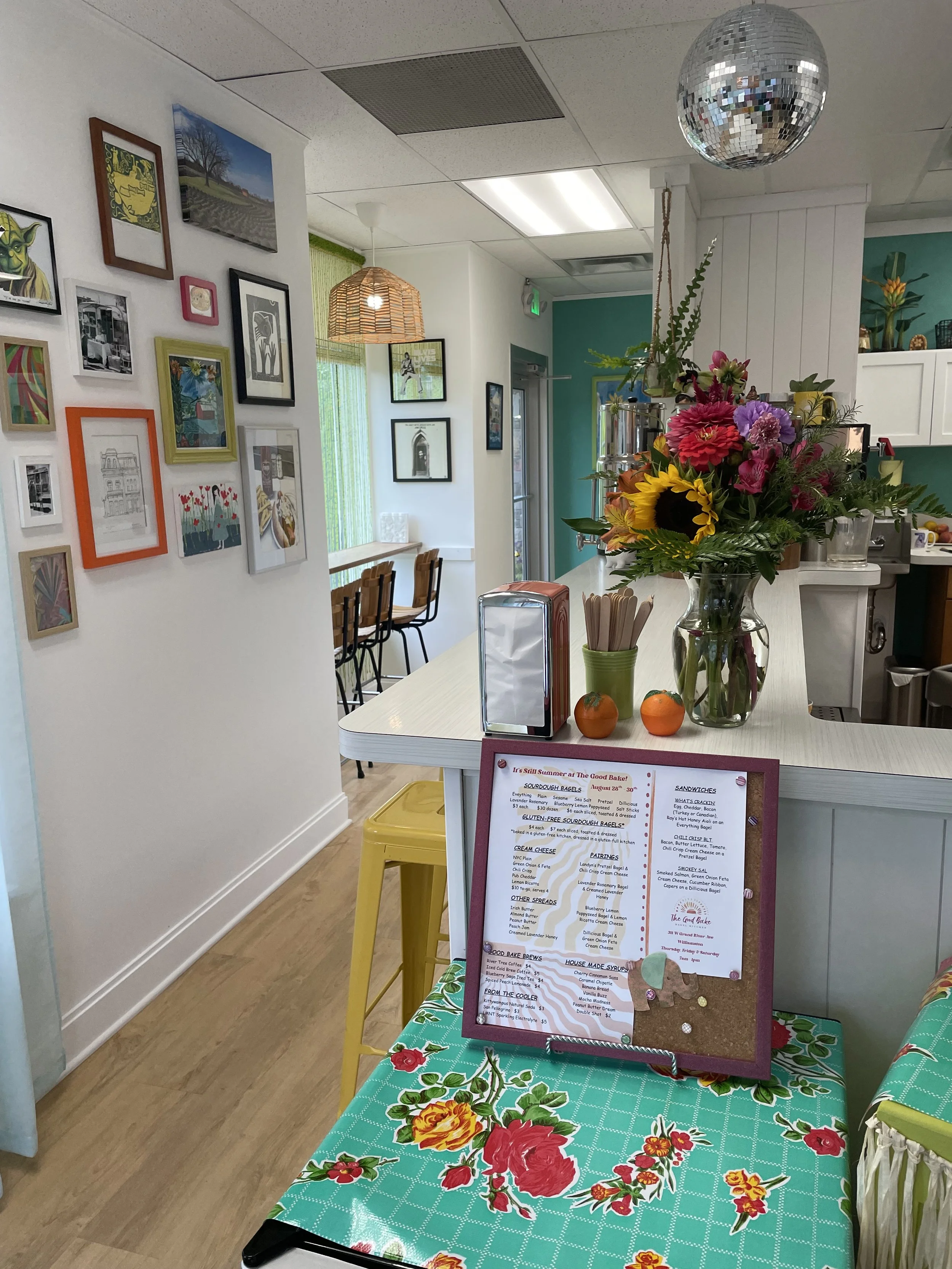 Interior of a cozy cafe with a stack of yellow stools, a bulletin board, a bouquet of colorful flowers, hanging disco ball, and wall art.