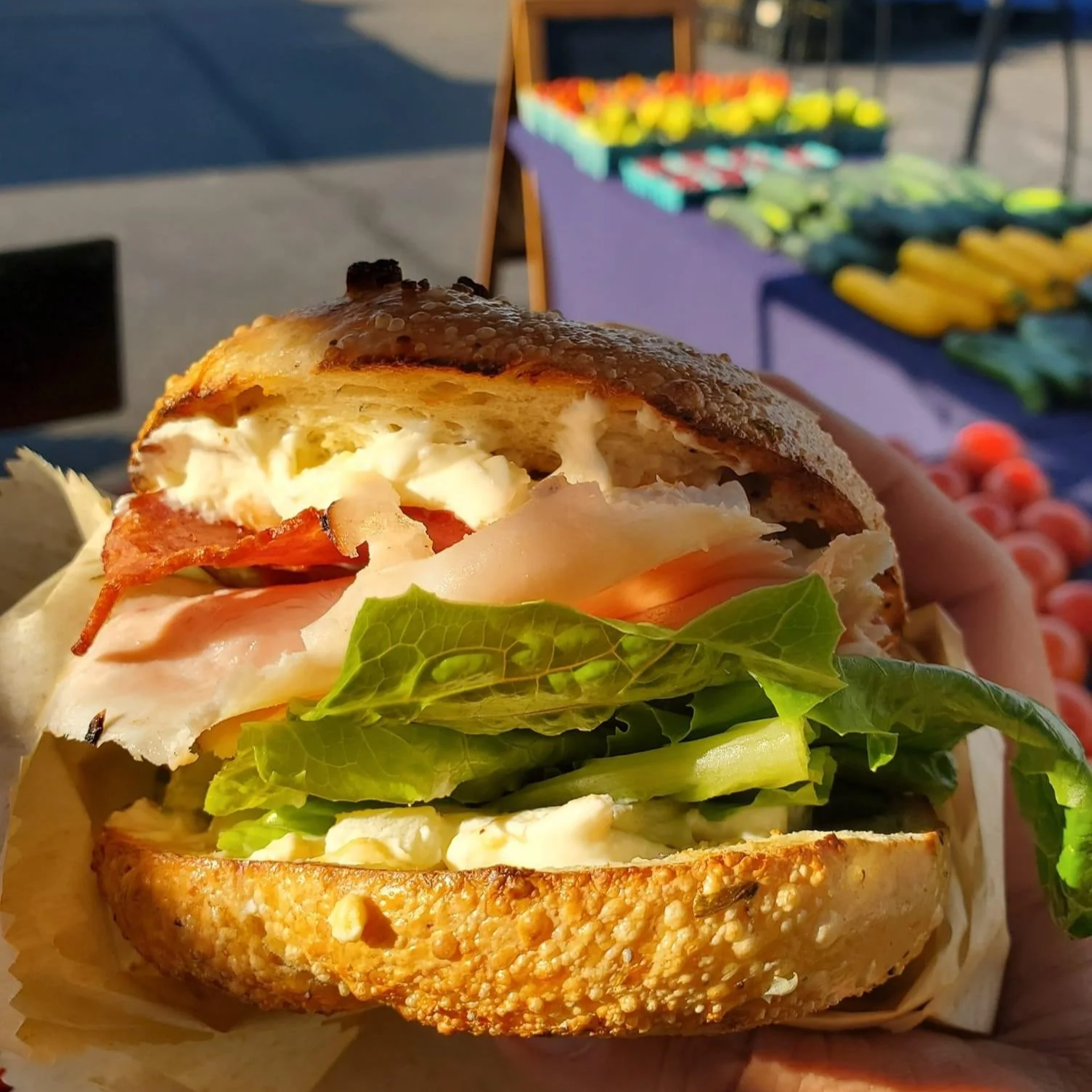 A close-up of a sandwich with lettuce, tomato, cheese, ham, bacon, egg, and a toasted bun, held outdoors near a local market or farm stand.