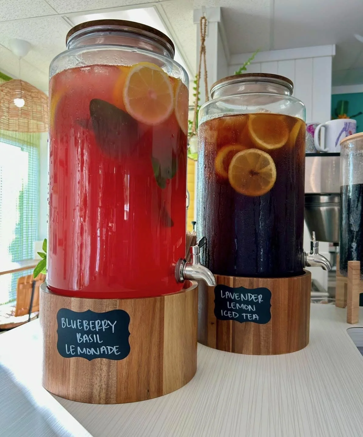 Two large glass beverage dispensers on a white table, one containing blueberry basil lemonade with lemon slices and blueberries, and the other containing lavender lemon iced tea with slices of lemon, both sitting on wooden bases with labels.
