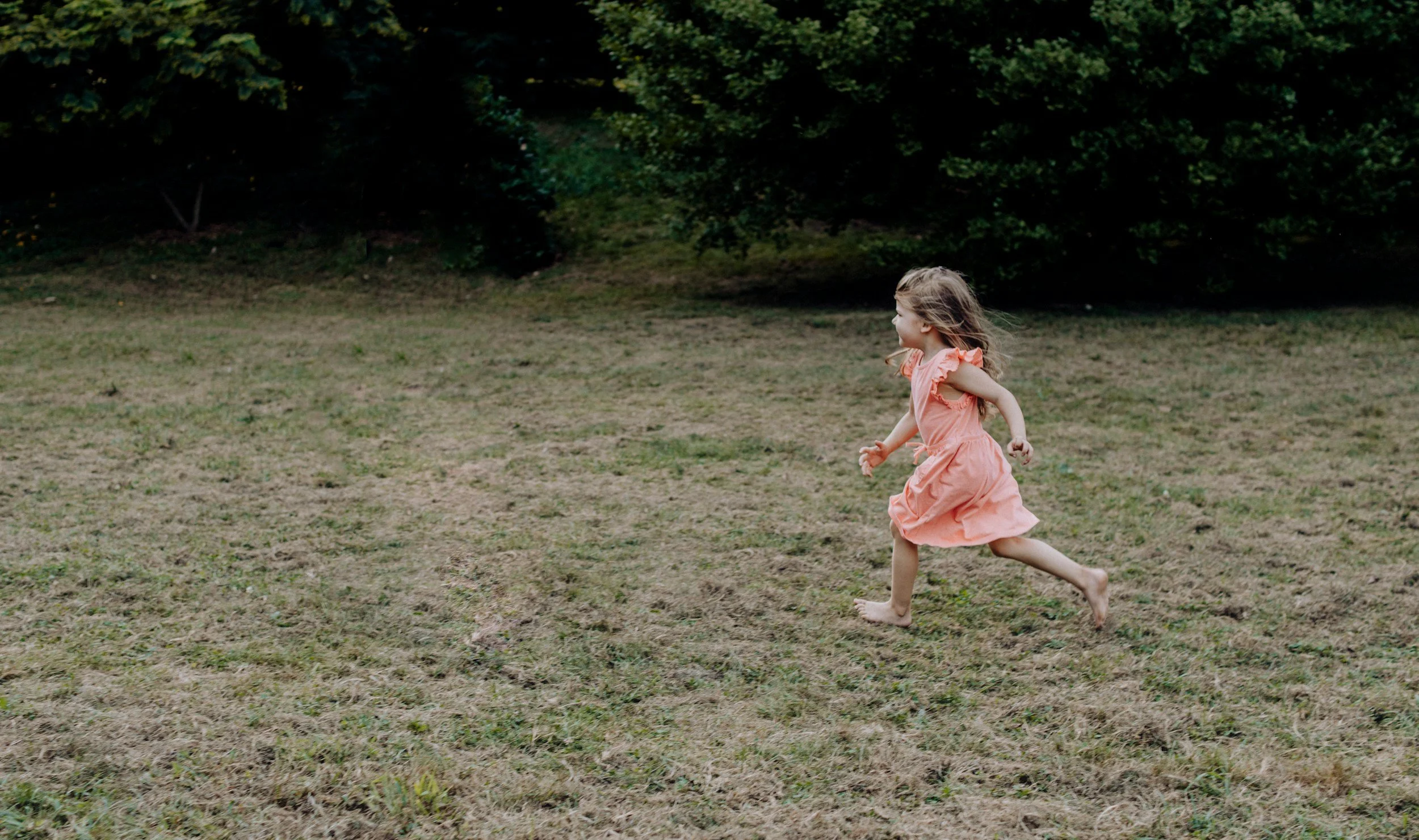 A young girl running barefoot across a grassy field with trees in the background, wearing a pink dress.