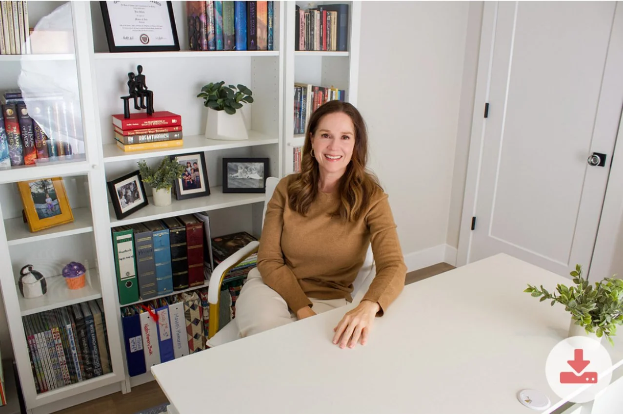 A woman with long wavy brown hair sitting at a white desk in a room with white walls and bookshelf behind her. The bookshelf holds books, framed photos, potted plants, and decorative items.