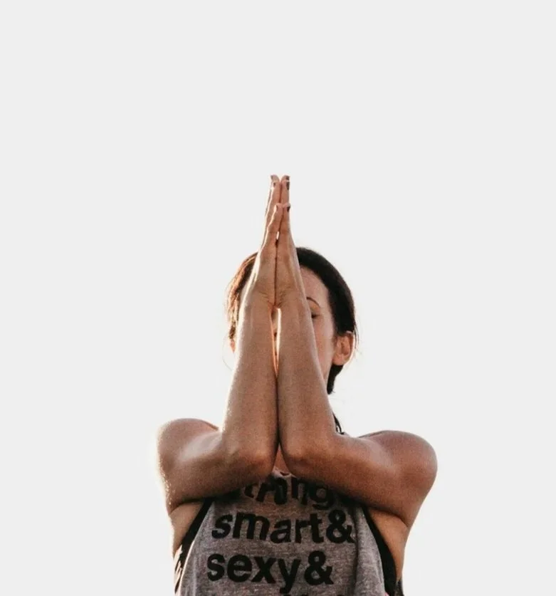 A woman with dark hair in a yoga pose with her palms pressed together in front of her face, against a plain white background.