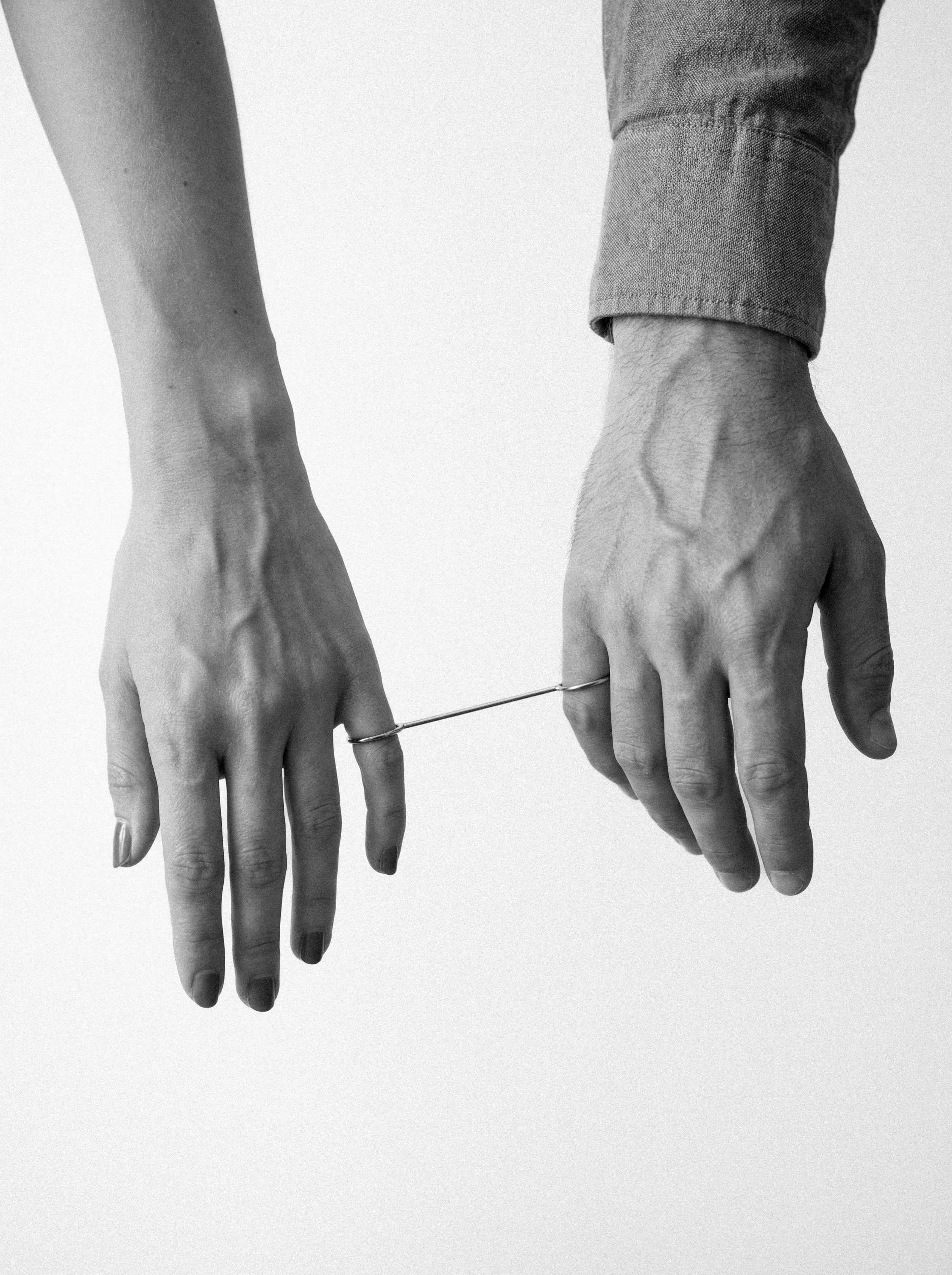 Black and white photograph of a person with dark painted nails holding a thin wire attached to another person's fingers. The person's hands are relaxed, and the background is plain.