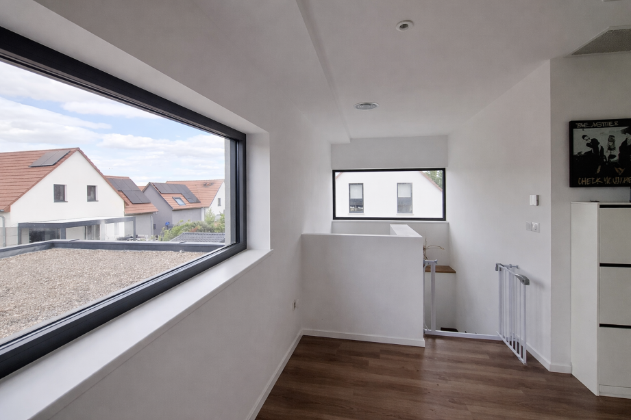 Vue intérieure d'une pièce avec deux fenêtres, murs blancs, sol en bois et quelques meubles et objets décoratifs.