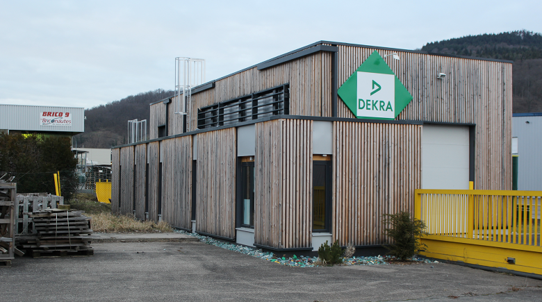 Bâtiment industriel avec façade en bois et logo Dekra, situé dans une zone avec d'autres bâtiments industriels, quelques plantes ornementales et un stockage de matériaux en extérieur.