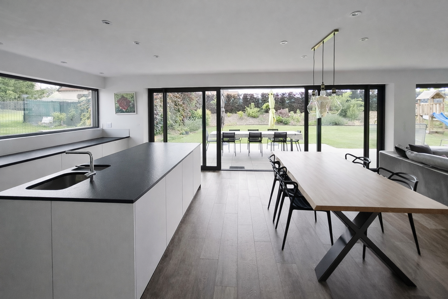 Cuisine moderne avec îlot central, grande table en bois avec six chaises, vue sur jardin extérieur avec patio, table de repas et aire de jeux pour enfants, luminosité naturelle