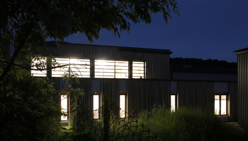 Bâtiment industriel avec fenêtres éclairées la nuit, entouré de végétation et de végétation, dans un environnement calme.