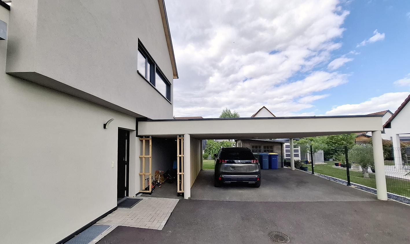 Un parking résidentiel avec une voiture noire, des poubelles bleues, un espace de stockage et un bâtiment résidentiel moderne en arrière-plan, sous un ciel partiellement nuageux.