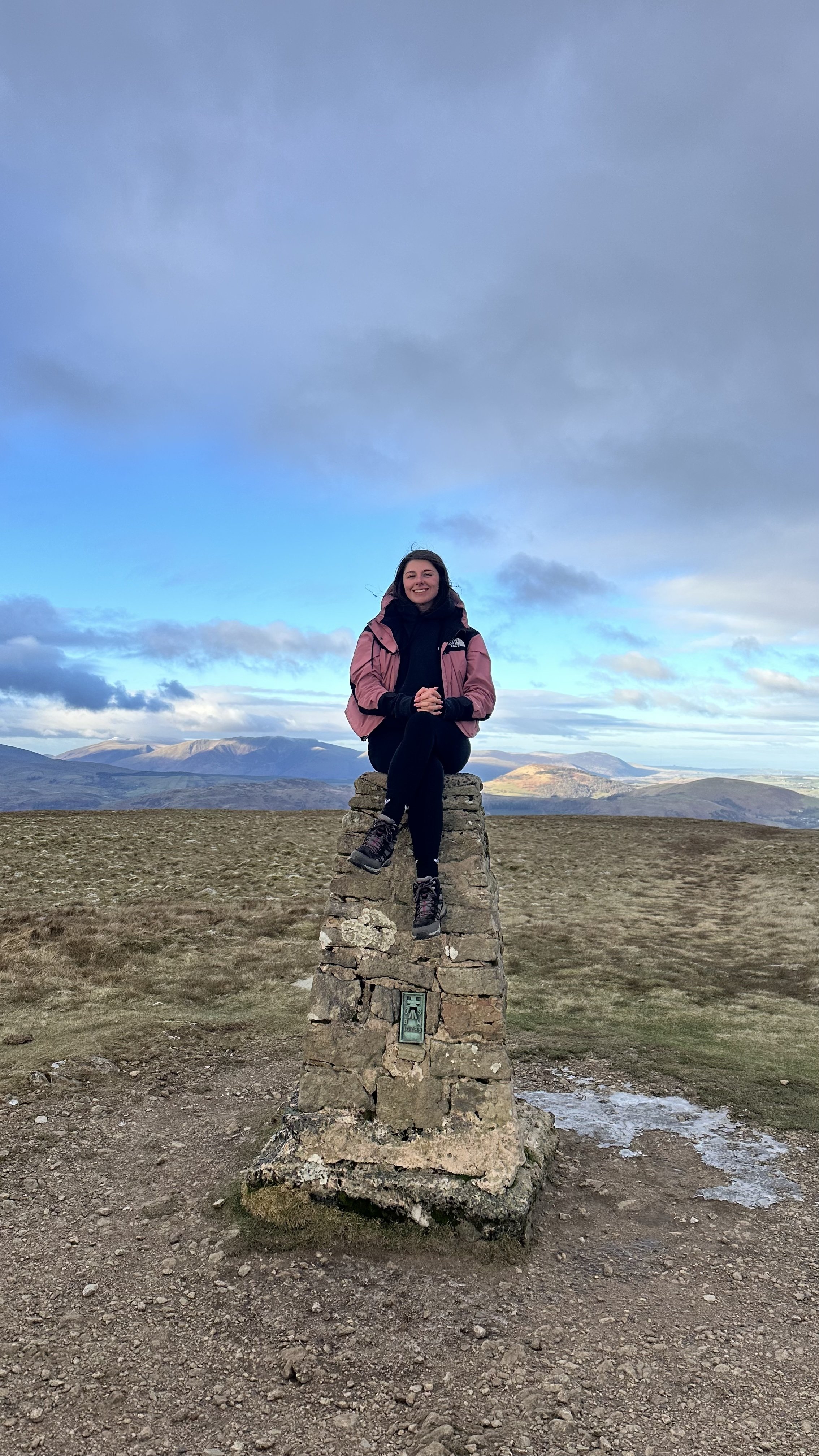 A woman sitting on top of a stone trig point marker on a hilltop with mountains in the background, under a mostly cloudy sky.