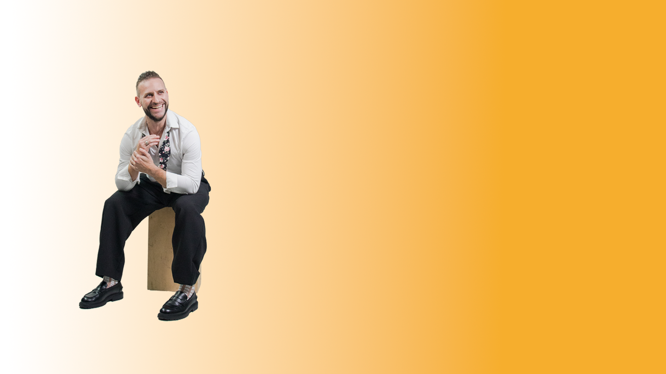 A man with a beard and short hair, wearing a white shirt and black pants, sitting on a wooden block, smiling and holding his hands together, with a yellow gradient background.