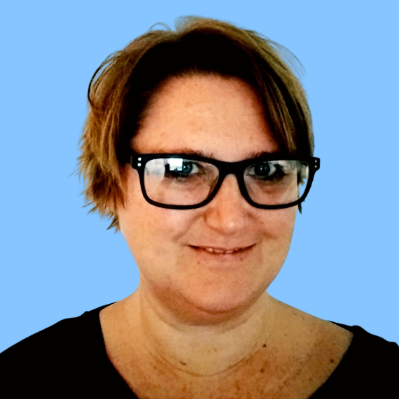 Portret van een vrouw met kort bruin haar en zwarte bril, lachend voor een lichtblauwe achtergrond.