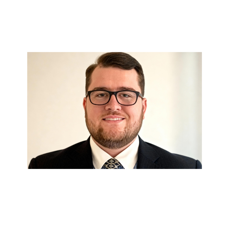 Portrait of a man with short brown hair, glasses, a beard, wearing a dark suit, white shirt, and patterned tie, smiling against a neutral background.